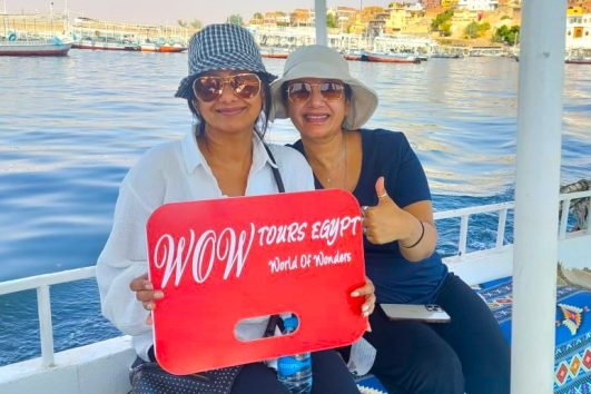 Two women sitting on a felucca boat on the Nile River in Aswan, holding a WOW Tours Egypt sign.