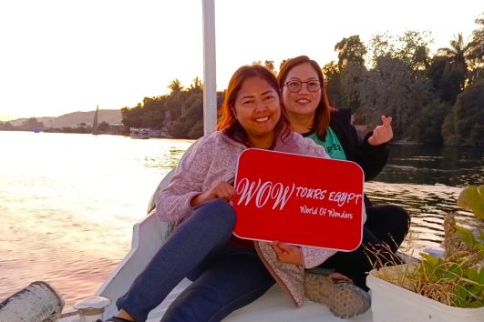 Two happy ladies enjoying a peaceful felucca boat ride on the Nile River at sunset, holding a WOW TOURS EGYPT sign. // Slide WOW // Aswan front (6)