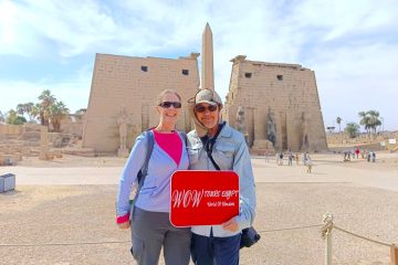Couple standing before the massive First Pylon and obelisk at Luxor Temple under a bright Egyptian sky.