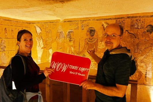 Two visitors holding a WOW Tours Egypt sign inside a decorated ancient Egyptian tomb in the Valley of the Kings, with wall paintings and hieroglyphs in the background // Aswan front (11)