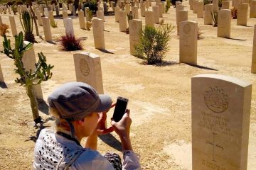 Visitor photographing a headstone at El Alamein War Cemetery in Egypt, with rows of graves and a memorial cross in the background