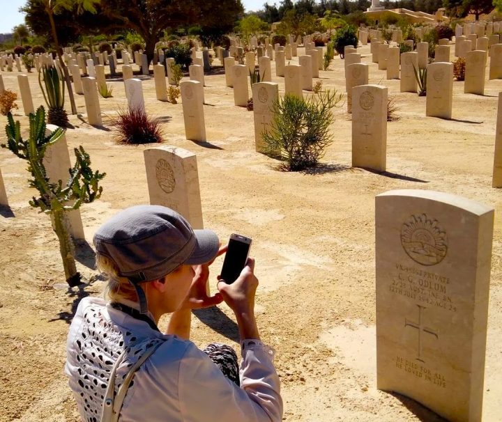 Visitor photographing a headstone at El Alamein War Cemetery in Egypt, with rows of graves and a memorial cross in the background