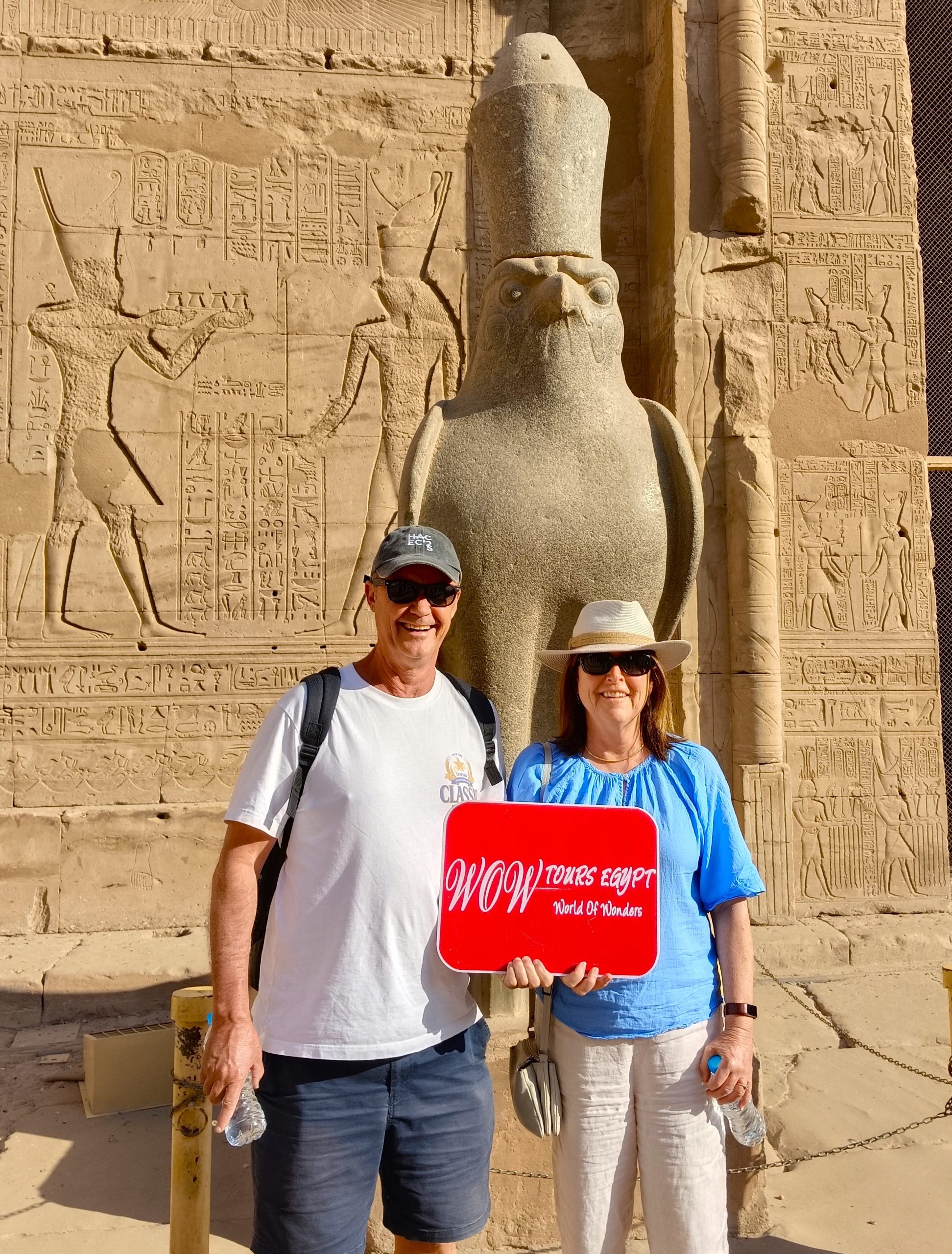 Happy couple standing in front of the falcon statue of Horus inside Edfu Temple holding the WOW Tours Egypt sign