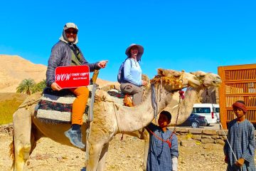Two guests riding camels on the west bank of Luxor, holding a WOW Tours Egypt sign, with local children, desert hills, and traditional buildings in the background