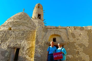 A couple stands in front of the ancient mud-brick architecture of Al-Qasr Islamic Village in Dakhla Oasis, holding WOW Tours Egypt sign.