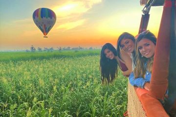 Three women looking out from a hot air balloon basket over green farmland in Luxor at sunrise, with another balloon flying in the distance