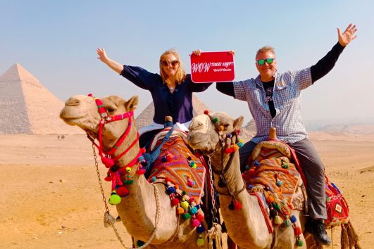 Couple riding camels at the Pyramids of Giza in Cairo with arms raised, holding a WOW TOURS EGYPT sign