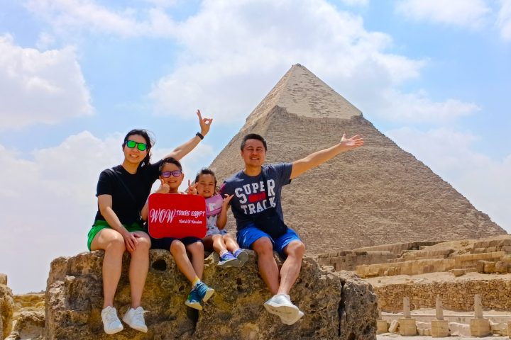 Family with two children sitting on rocks holding WOW TOURS EGYPT sign at the Great Pyramids of Giza in Cairo.