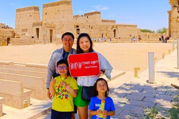 a smiling Family visiting Philae Temple in Aswan, Egypt, holding WOW TOURS EGYPT sign in front of Isis Temple during a private guided tour