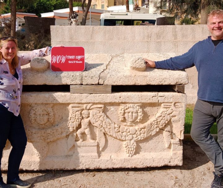 Couple holding WOW Tours Egypt sign beside a stone sarcophagus in the open area of the Catacombs of Kom El Shoqafa in Alexandria