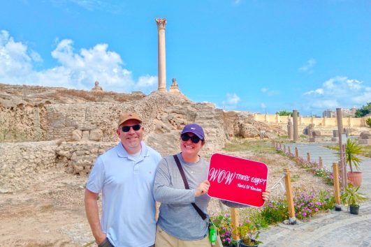 Happy guests standing at the historic Pompey's Pillar in Alexandria, holding WOW TOURS EGYPT sign.