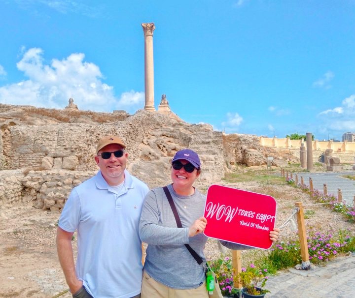 Happy guests standing at the historic Pompey's Pillar in Alexandria, holding WOW TOURS EGYPT sign.