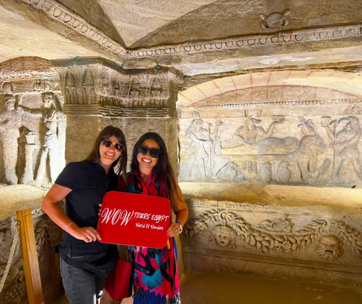 Two female travelers inside the Catacombs of Kom El Shoqafa beside an elaborately carved burial chamber in Alexandria.