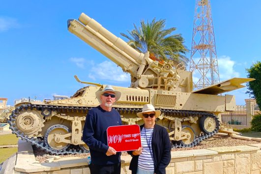 A couple standing beside a restored World War II artillery vehicle in El Alamein, Egypt during a historical guided tour.