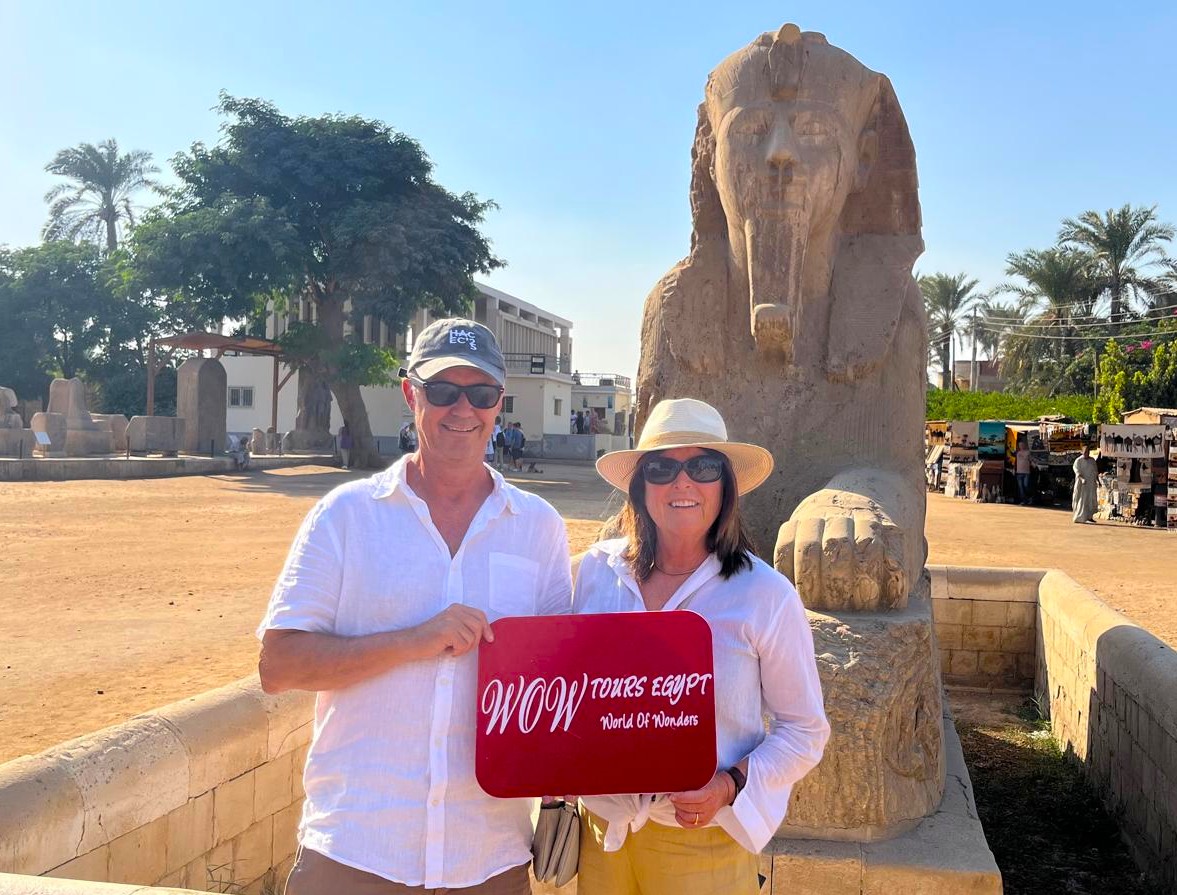A couple holding a WOW TOURS EGYPT sign in front of the alabaster Sphinx at the Memphis Open-Air Museum in Egypt.